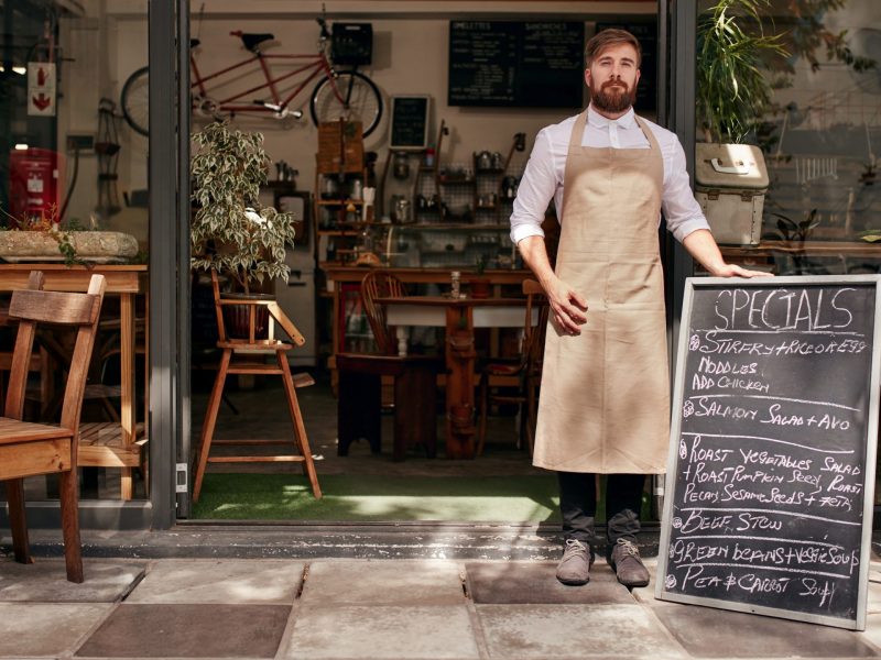 Portrait of young man standing at the entrance of his coffee shop with a black board. Barista standing in the doorway of a restaurant.